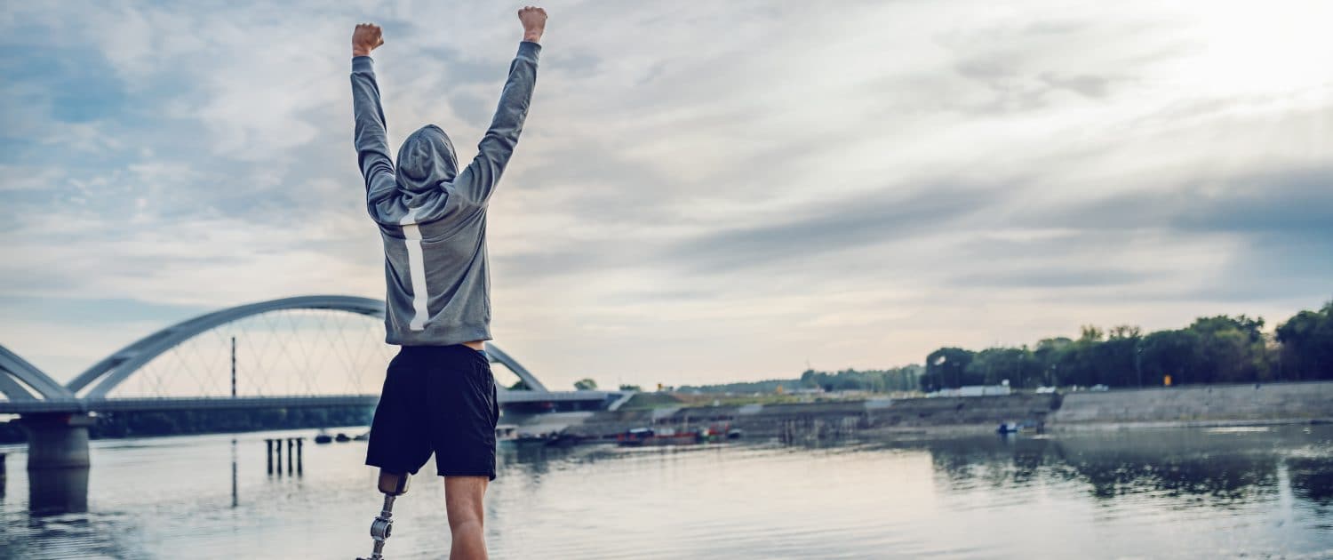 Highly motivated caucasian handicapped sportsman in sportswear and with artificial leg standing on quay and holding arms up. Only way to win is to be yourself.